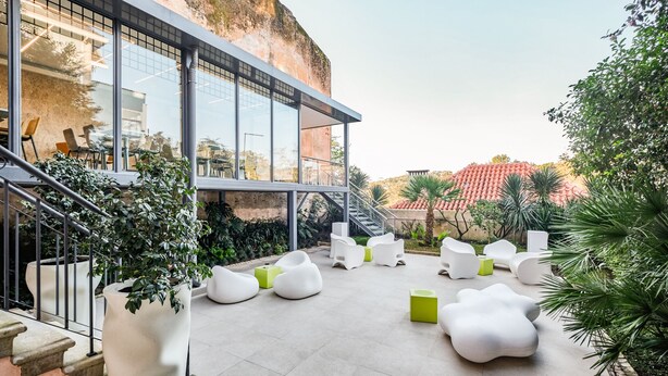 A modern outdoor seating area with white and green chairs, large potted plants, and a staircase leading to a glass building.