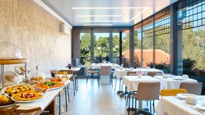 A dining area with tables set for breakfast, a buffet with various food items, and large windows offering a view of the outdoors.
