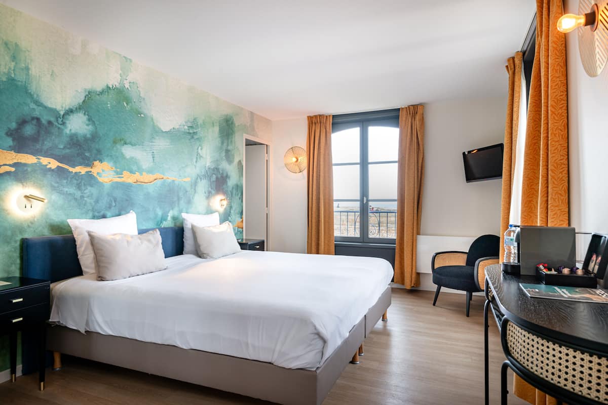 A modern hotel room with a large bed, a desk with a laptop, a chair, and a view of the outdoors through a window.