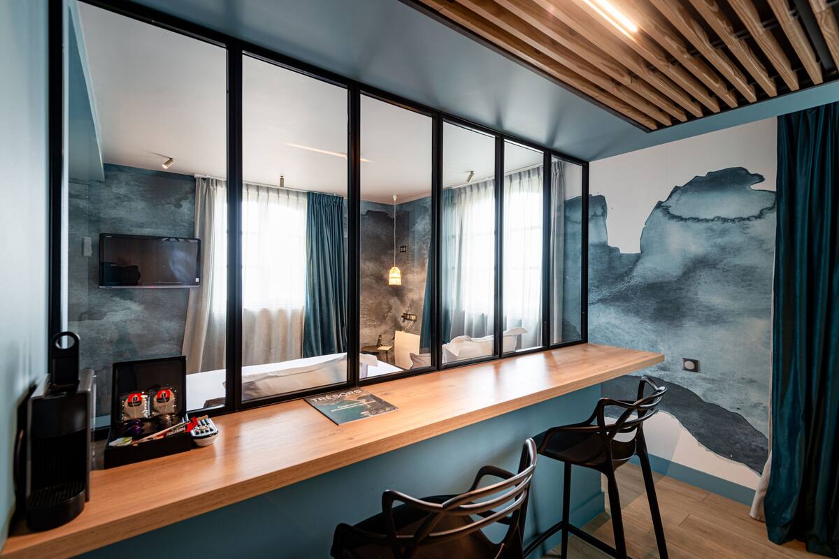 A modern room with a wooden desk, two bar stools, a large mirror, and a view of a bedroom through the window.
