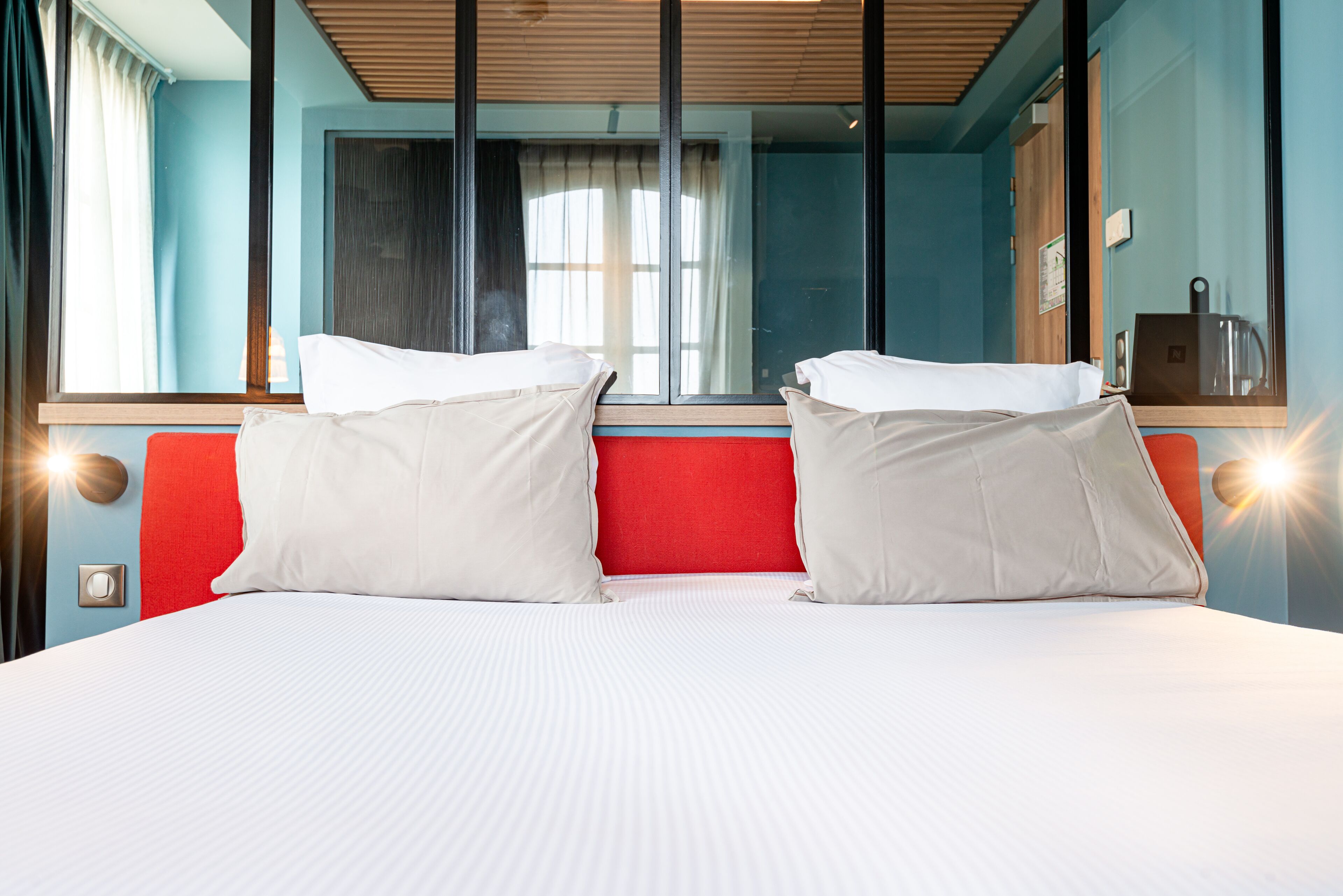 Cama doble con ropa de cama blanca y dos almohadas blancas, cabecera roja y una ventana grande con cortinas.