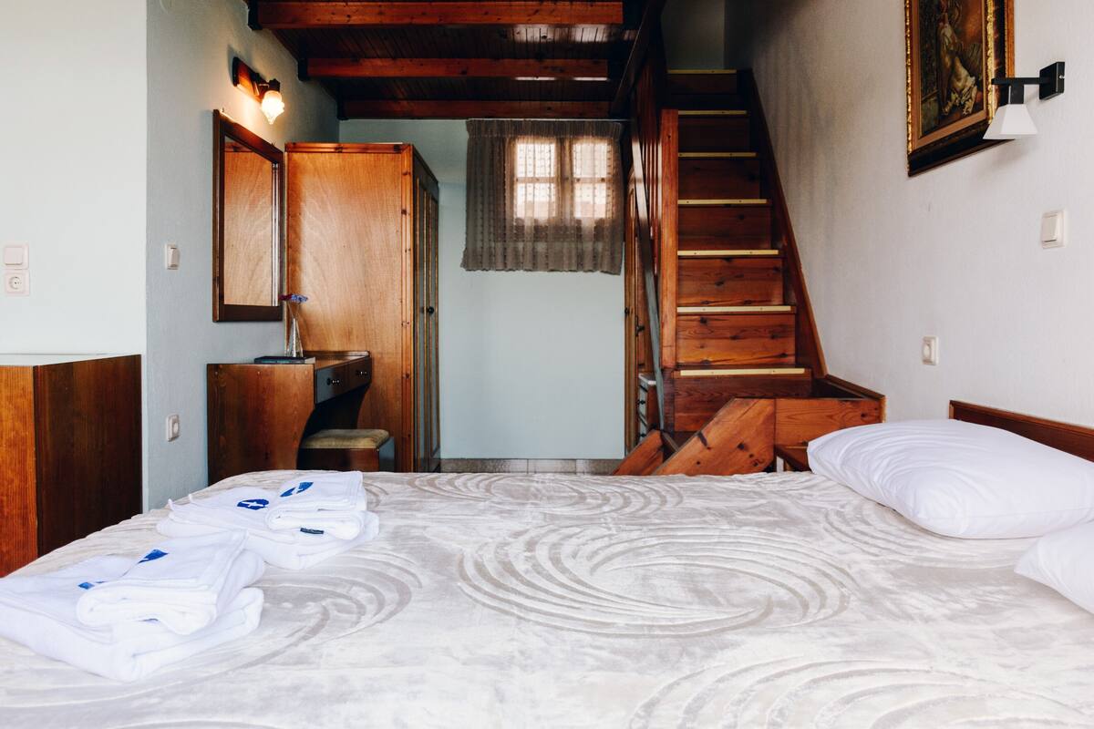 Un dormitorio con una cama, un aparador de madera, un espejo, una ventana con cortinas y una escalera.
