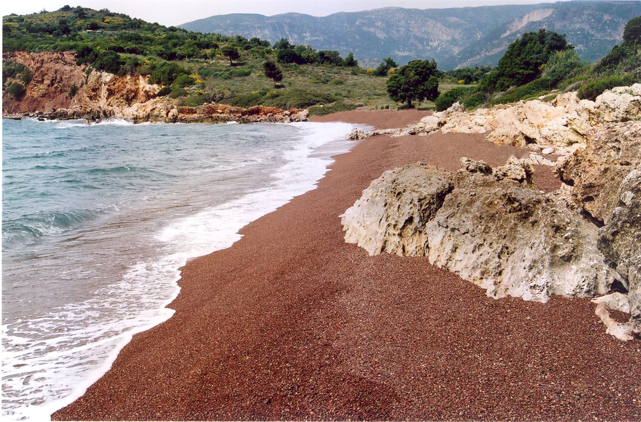 Ein Strand mit roten Kieselsteinen, felsigen Klippen und grünen Hügeln im Hintergrund.
