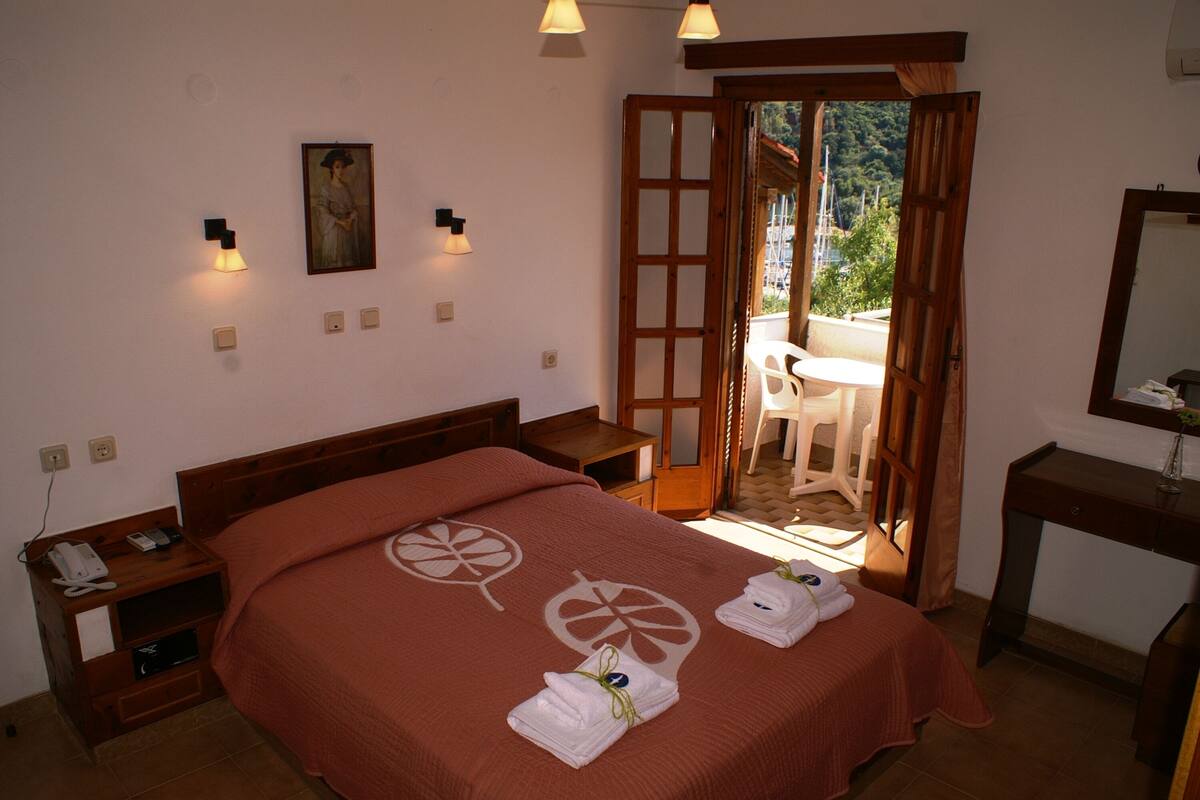 Habitación de hotel con cama, mesita de noche y escritorio con silla. Se ve una puerta que da a un espacio exterior con mesa y sillas.
