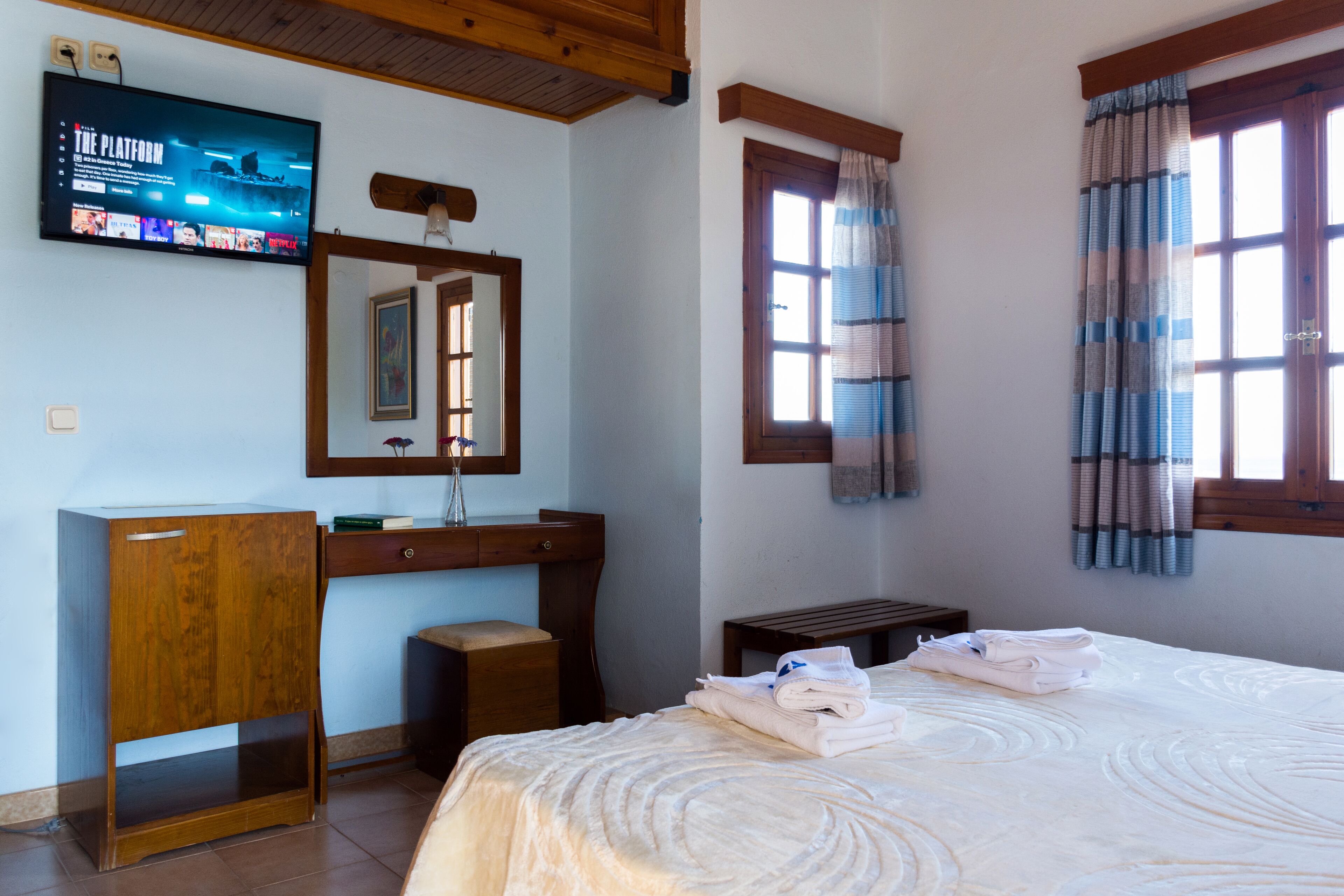 A hotel room with a bed, a wooden desk, a television mounted on the wall, and a window with curtains.