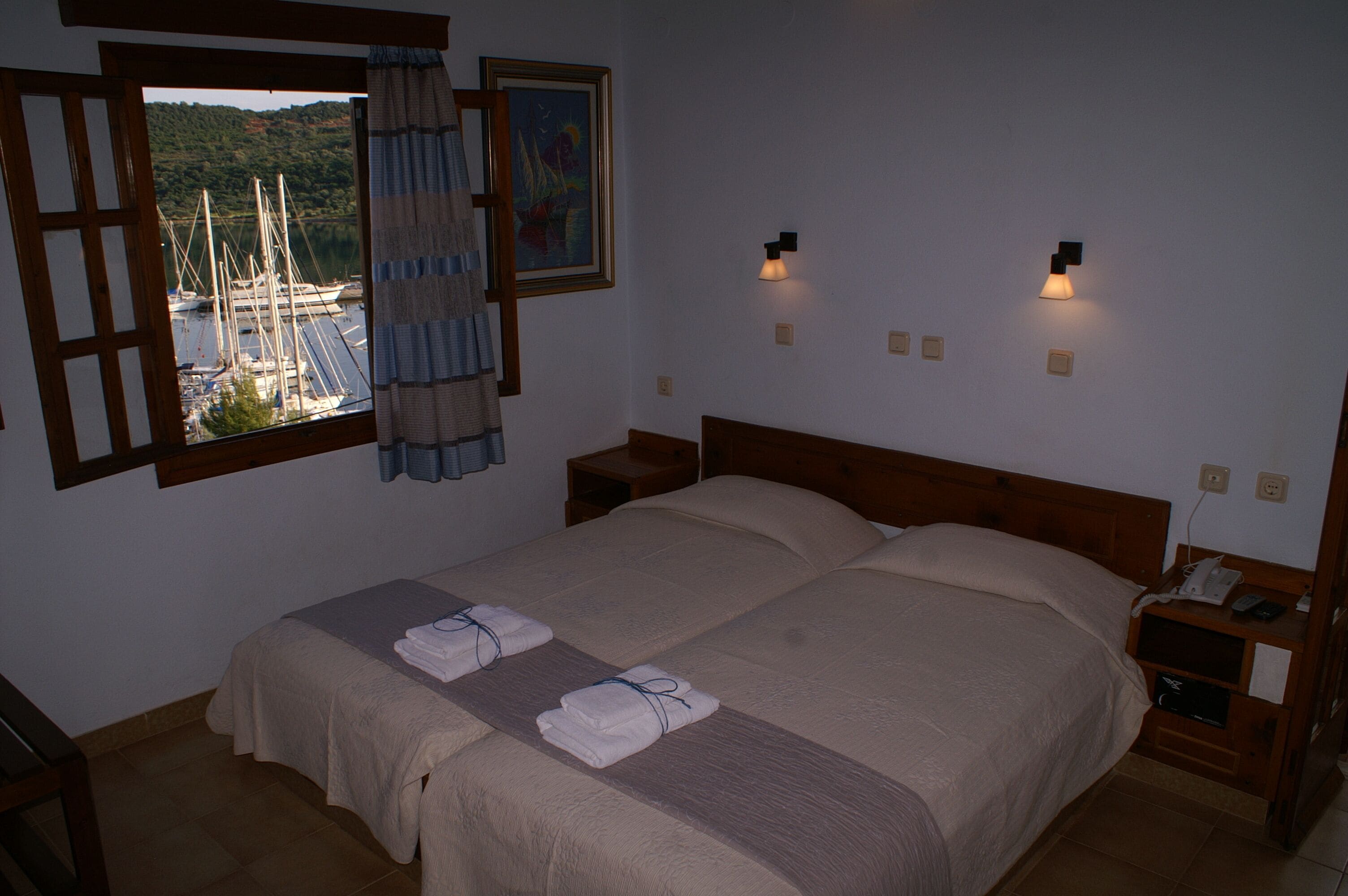 Una cama doble con dos almohadas, una mesita de noche con lámpara, una ventana con cortinas y un cuadro en la pared.