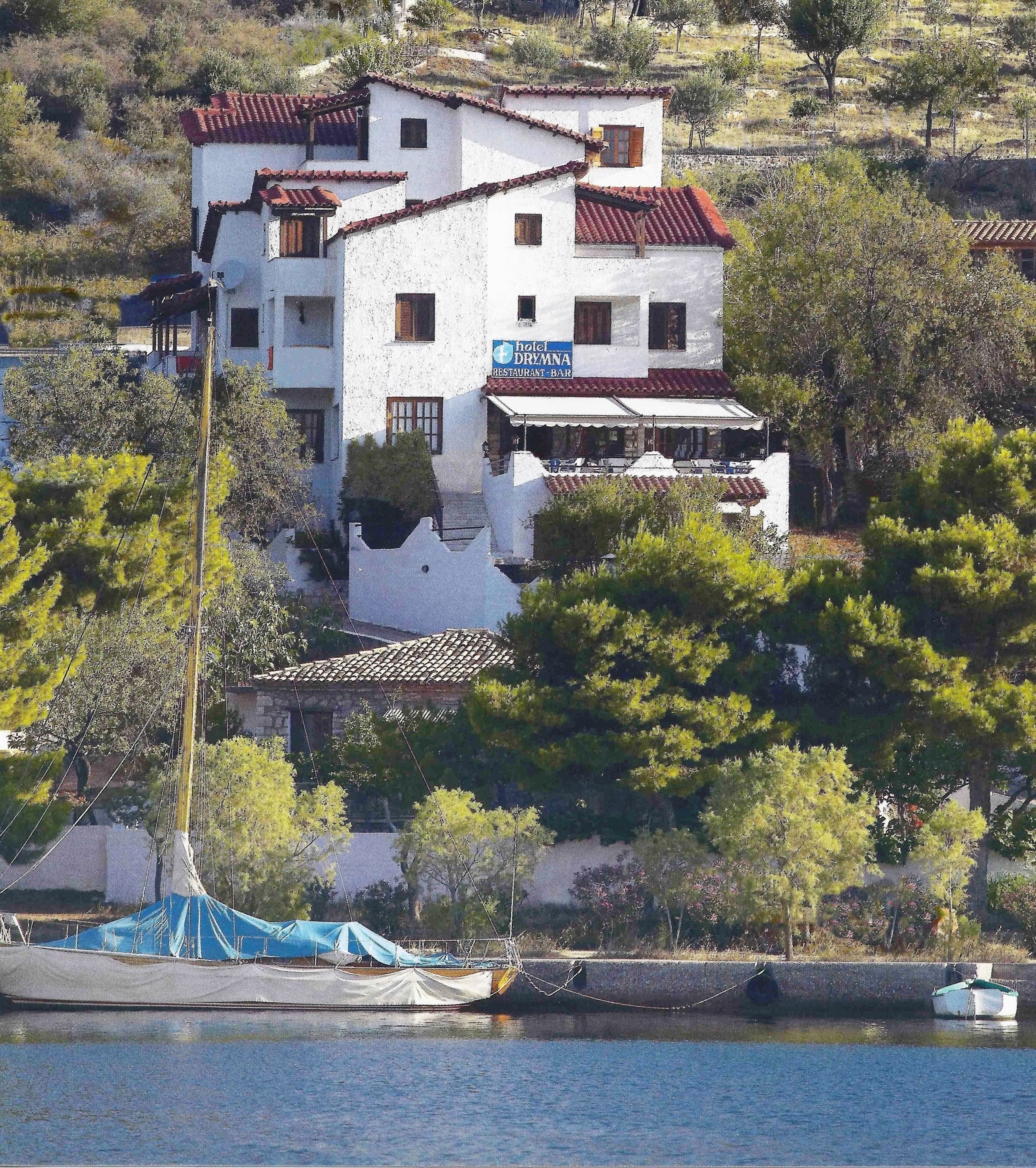 Un bâtiment blanc avec des toits recouverts de tuiles rouges, une enseigne indiquant « Hôtel Dremna, restaurant-bar », et un bateau amarré au bord de l’eau.