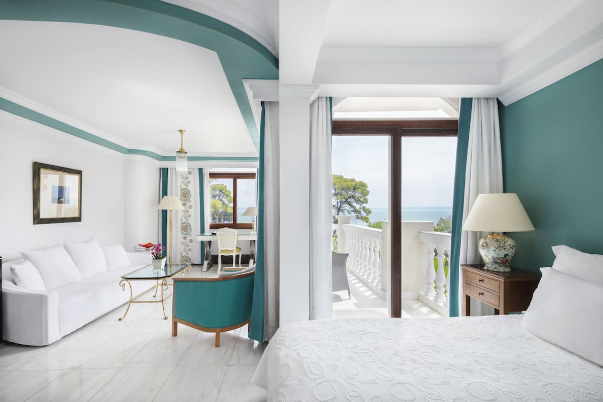 Een hotelkamer met een bed, een bank, een bureau en een balkon met uitzicht op zee.