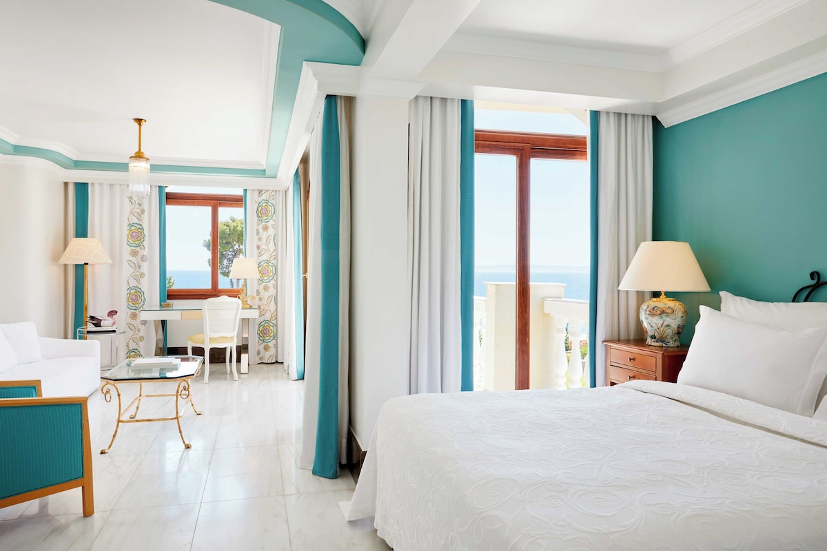 A hotel room with a large bed, a desk, a chair, and a view of the sea.