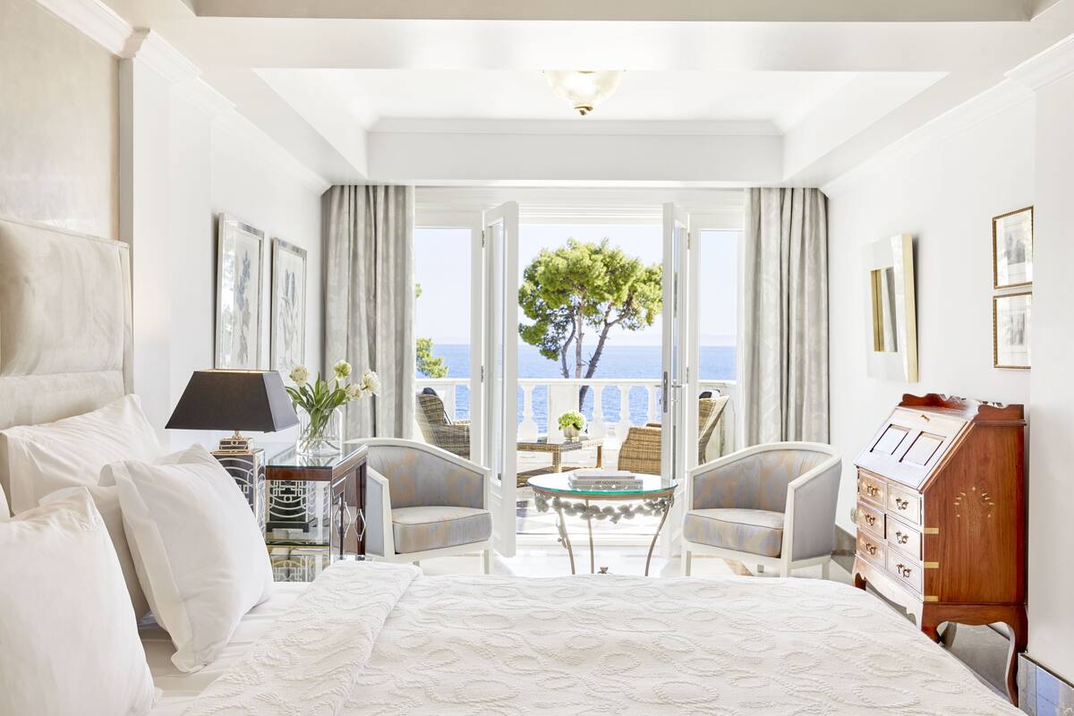 Hotelkamer met een groot bed, twee fauteuils, een klein tafeltje en een dressingkast. De kamer heeft een balkon met uitzicht op zee en bomen.