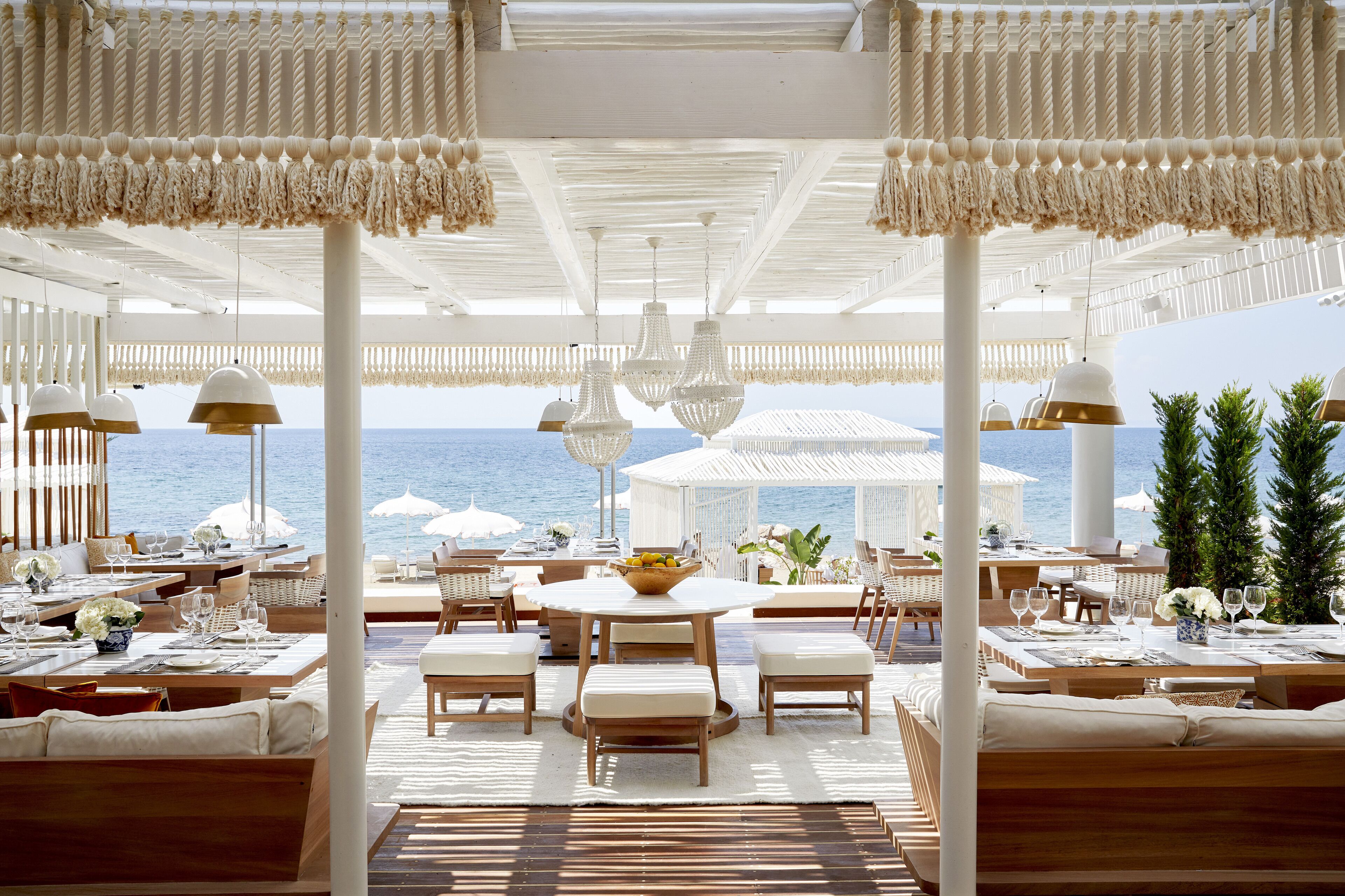 A luxurious outdoor dining area with white furniture, hanging chandeliers, and a view of the ocean.