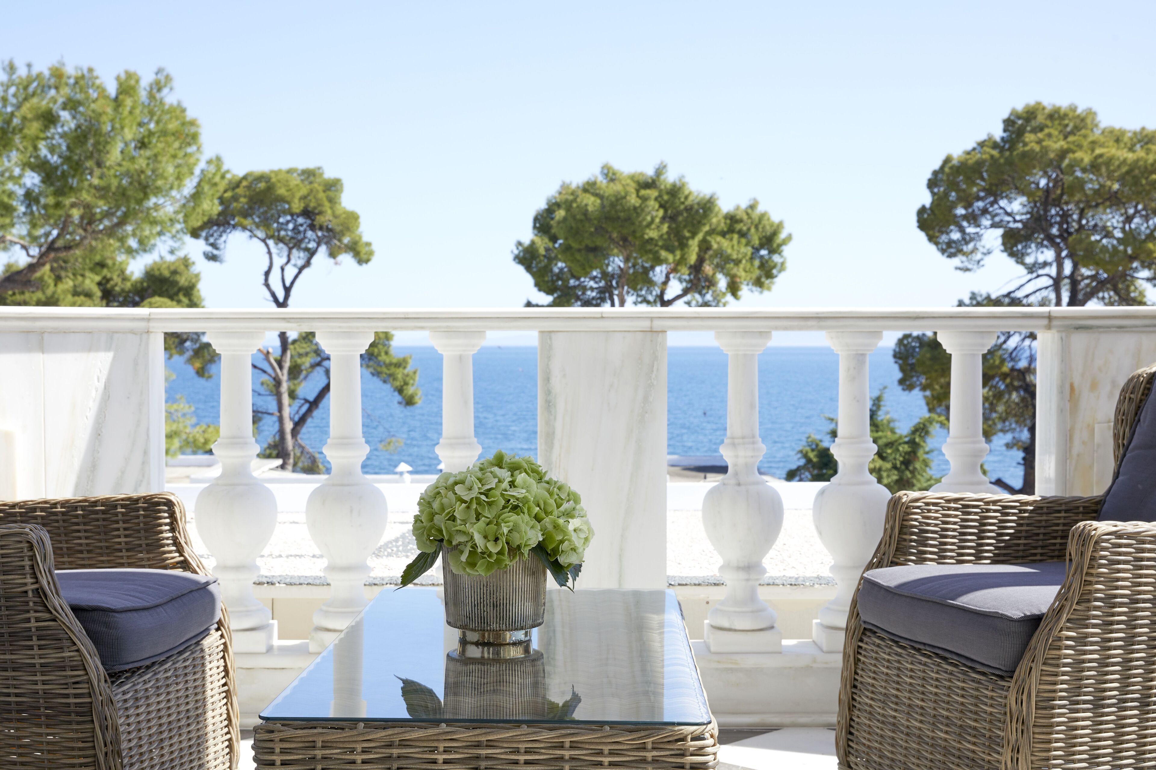 Un balcón con muebles de mimbre y una mesa, con vistas a un mar azul cristalino y árboles.
