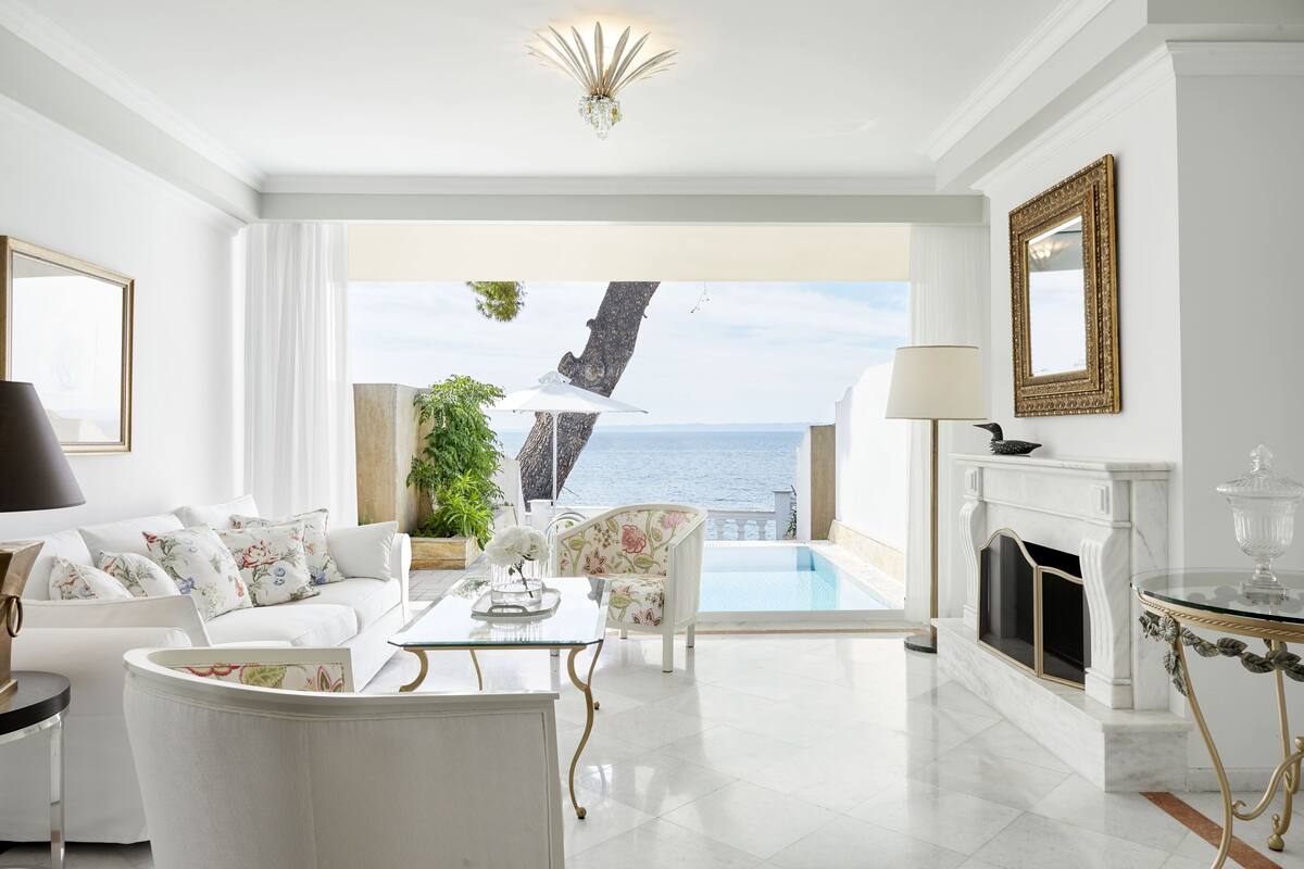 Een ruime woonkamer met een wit kleurenschema, een open haard, een glazen tafel en uitzicht op zee.