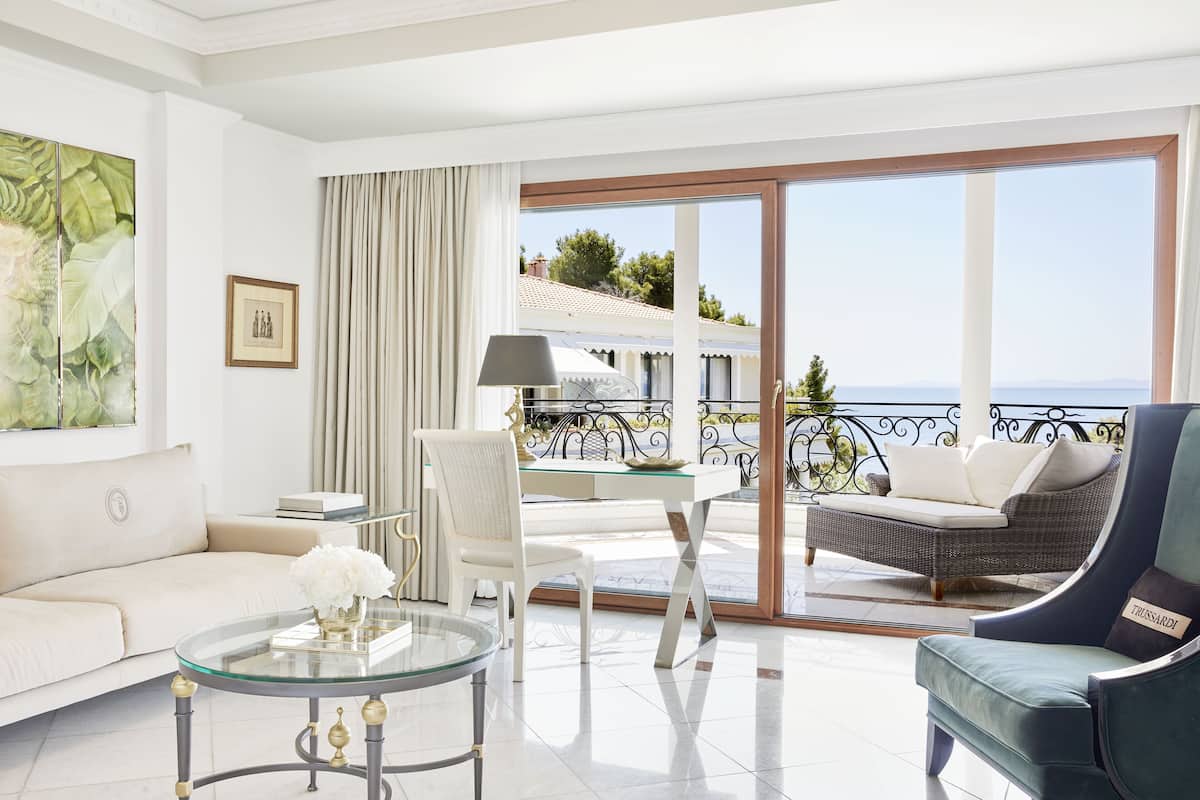 Een moderne woonkamer met een glazen salontafel, een witte bank en een donkere fauteuil. Een eettafel met een lamp en stoel is zichtbaar. De kamer heeft een grote glazen schuindeur die leidt naar een balkon met uitzicht op zee en gebouwen.