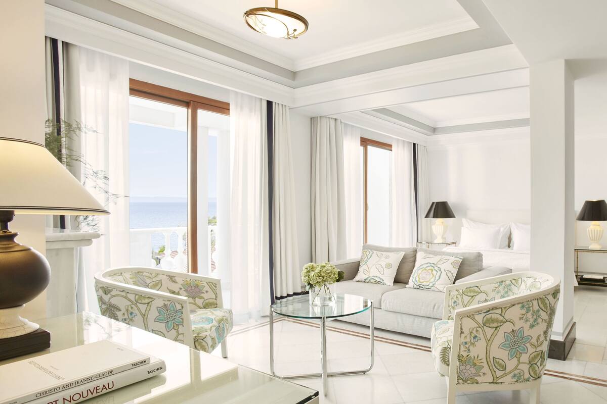Een ruime woonkamer met een bank, fauteuils, een glazen salontafel en uitzicht op zee door grote ramen.