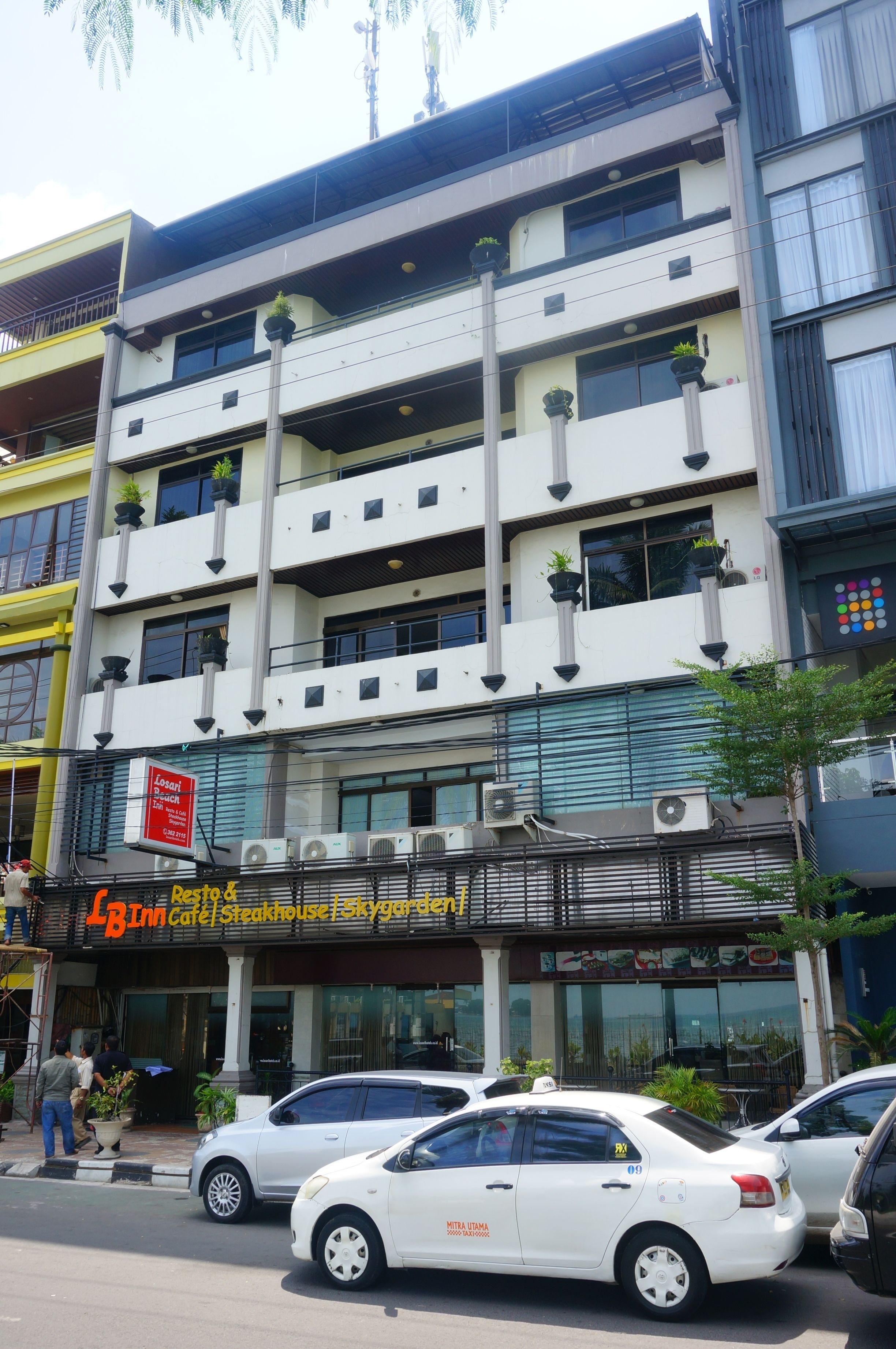A multi-story building with a restaurant on the ground floor, multiple balconies with plants, and parked cars on the street.