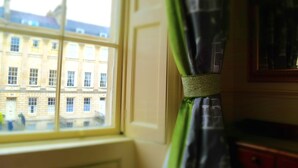 A room with a window overlooking a building, a curtain, and a framed picture on the wall. - The Windsor Townhouse (Bath)