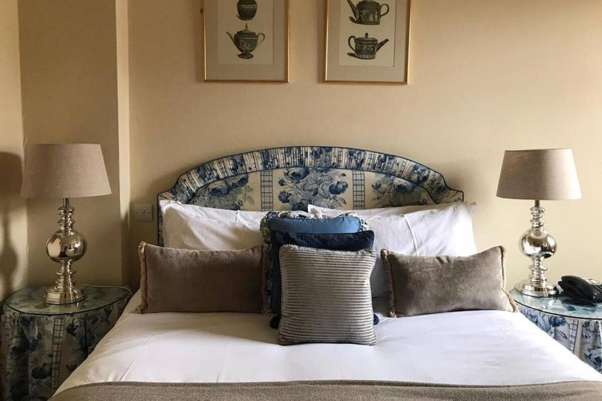 Una cama bien hecha con cabecera azul y blanca, dos mesitas de noche con lámparas, y dos cuadros enmarcados en la pared.