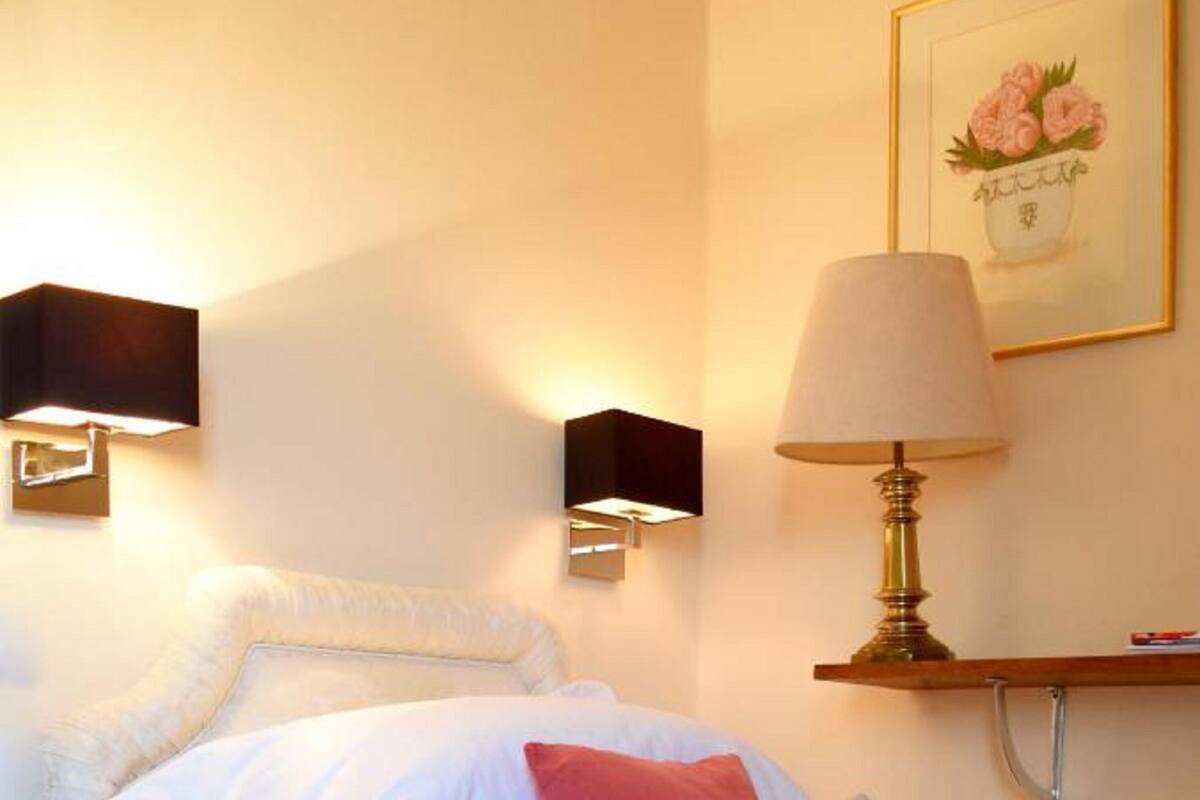 Una habitación de hotel con una cama, almohadas, una lámpara fijada a la pared y un cuadro enmarcado.