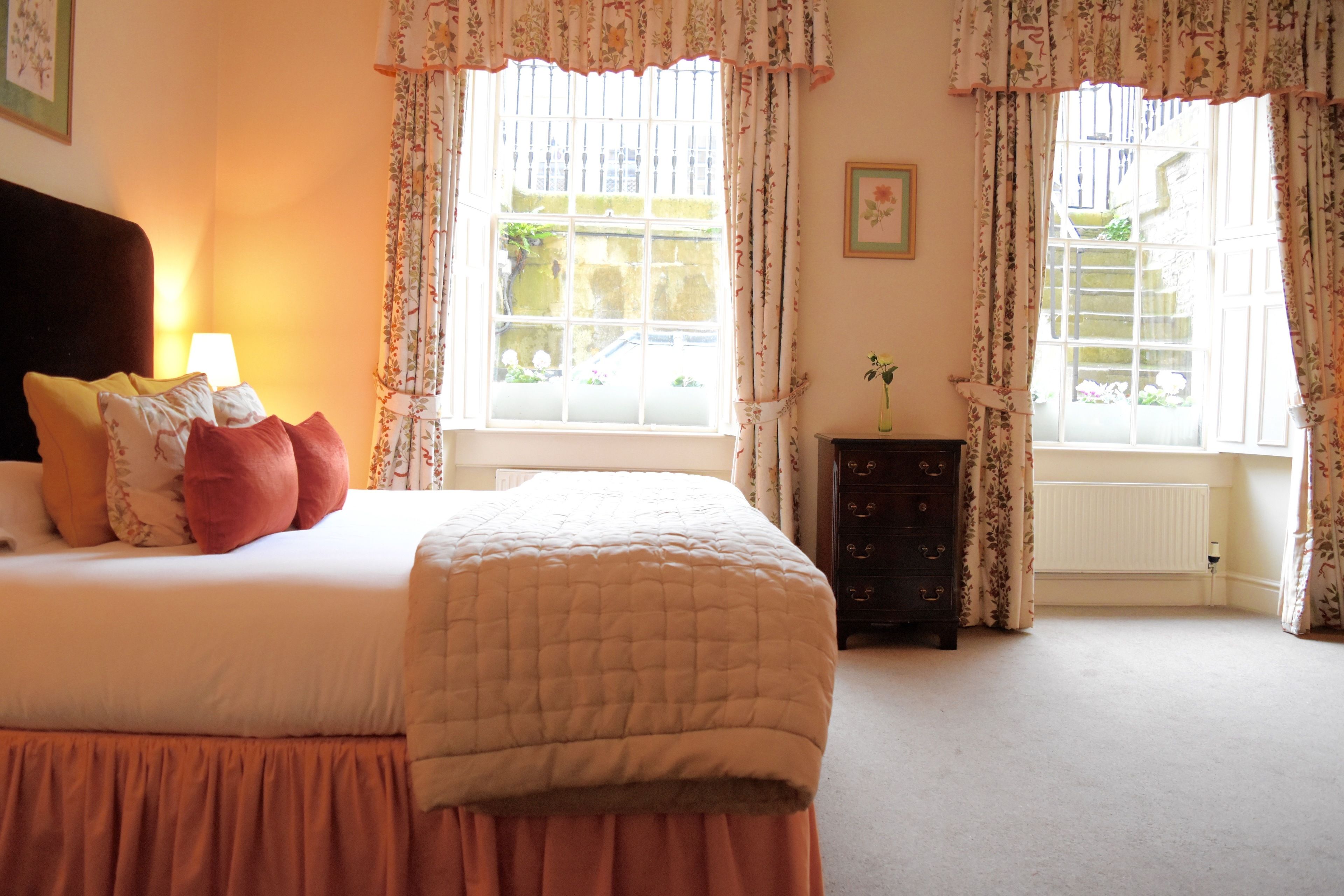 A bedroom with a bed, pillows, a dresser, and two windows with curtains.