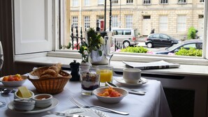 A breakfast table with various dishes, a basket of bread, and a window view of a city street. - The Windsor Townhouse (Bath)