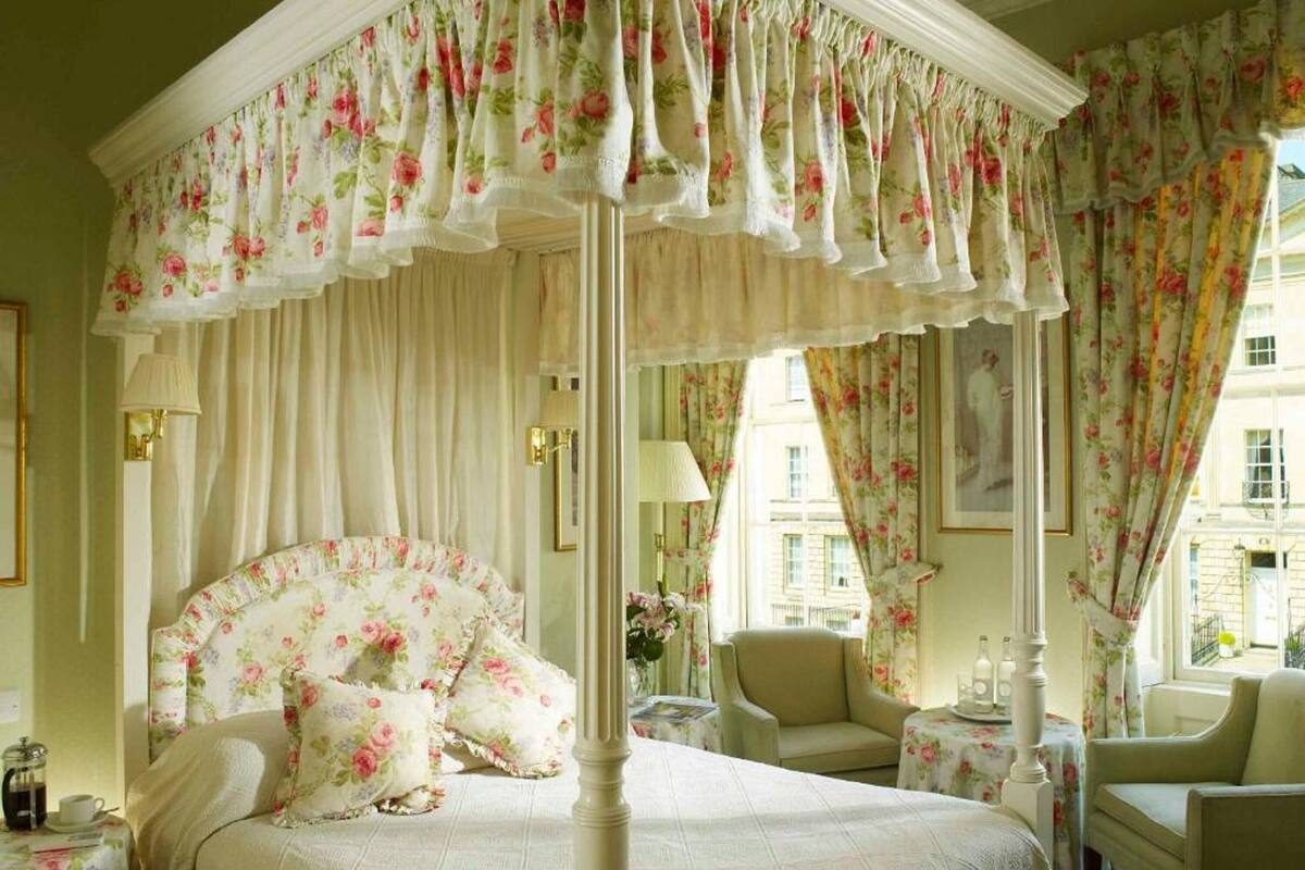 Una cama de estilo clásico con dosel y cortinas florales, un área de estar con una mesa y una silla, y vistas de la ciudad a través de grandes ventanales.