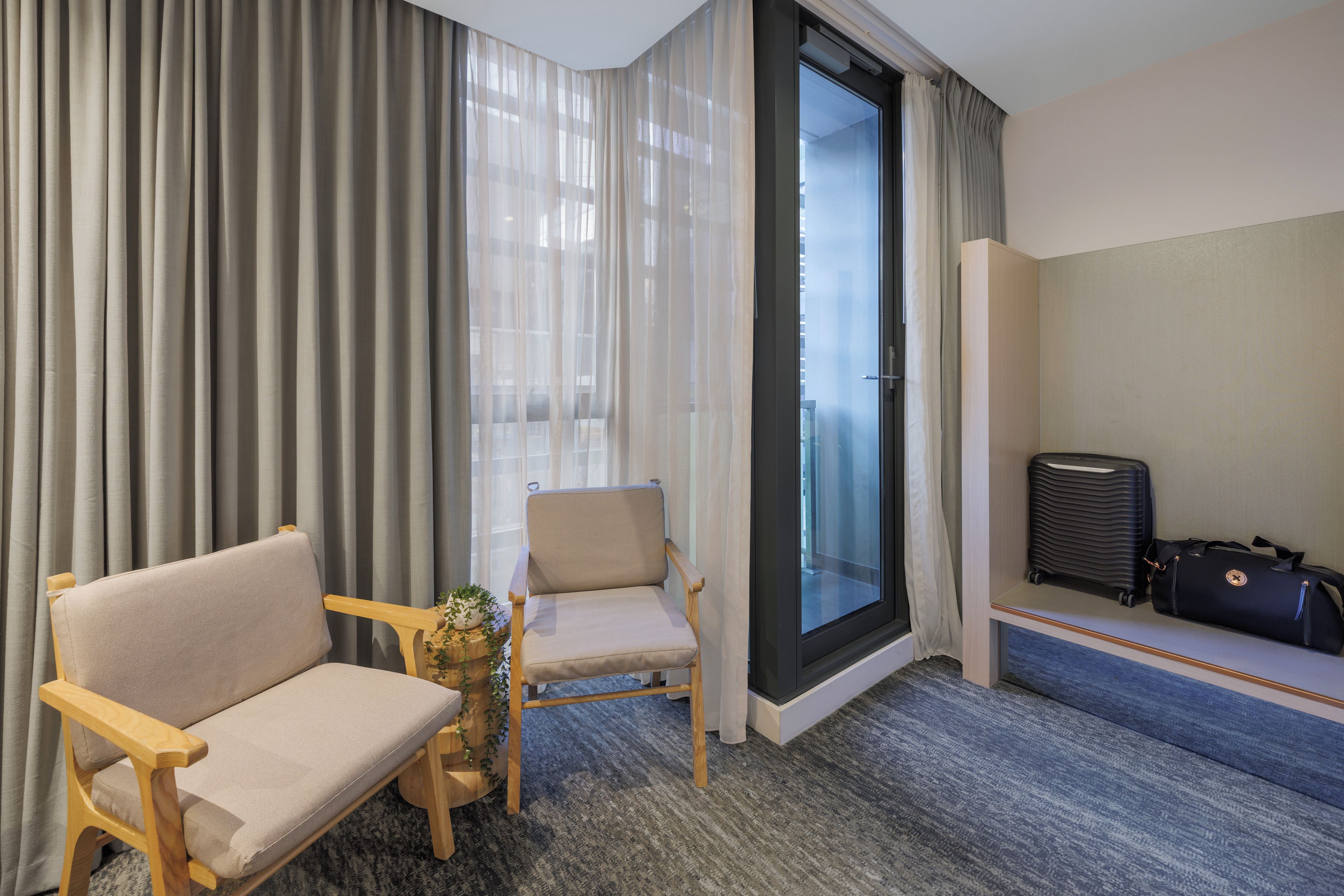 Une chambre d’hôtel avec un canapé, un fauteuil, une petite table avec une plante, une valise et une porte-fenêtre coulissante donnant sur un balcon.