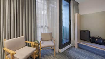 Une chambre d’hôtel avec un canapé, un fauteuil, une petite table avec une plante, une valise et une porte-fenêtre coulissante donnant sur un balcon.