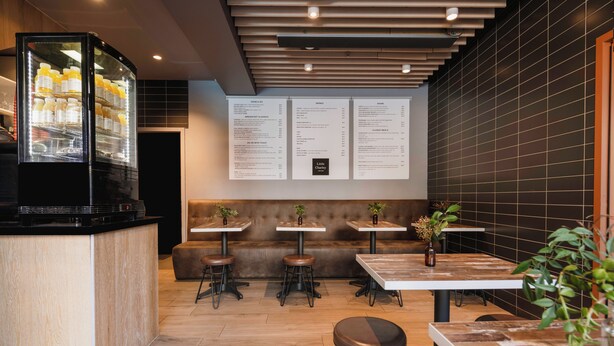 Interior de una cafetería moderna con mostrador, zona de asientos, carteleras de menú y un refrigerador.