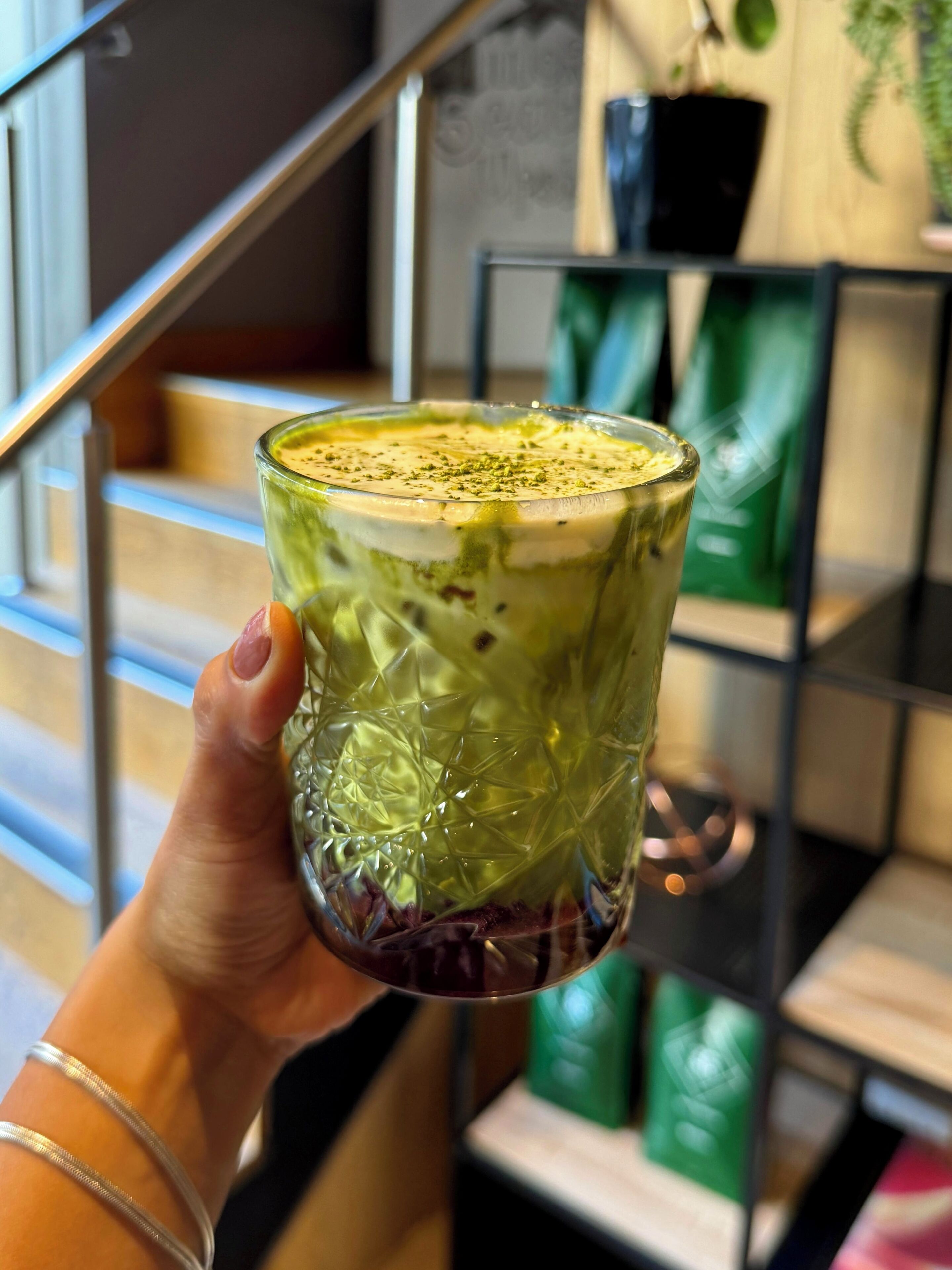 A hand holding a glass with a layered beverage, a metal staircase, and a shelf with green items in the background.