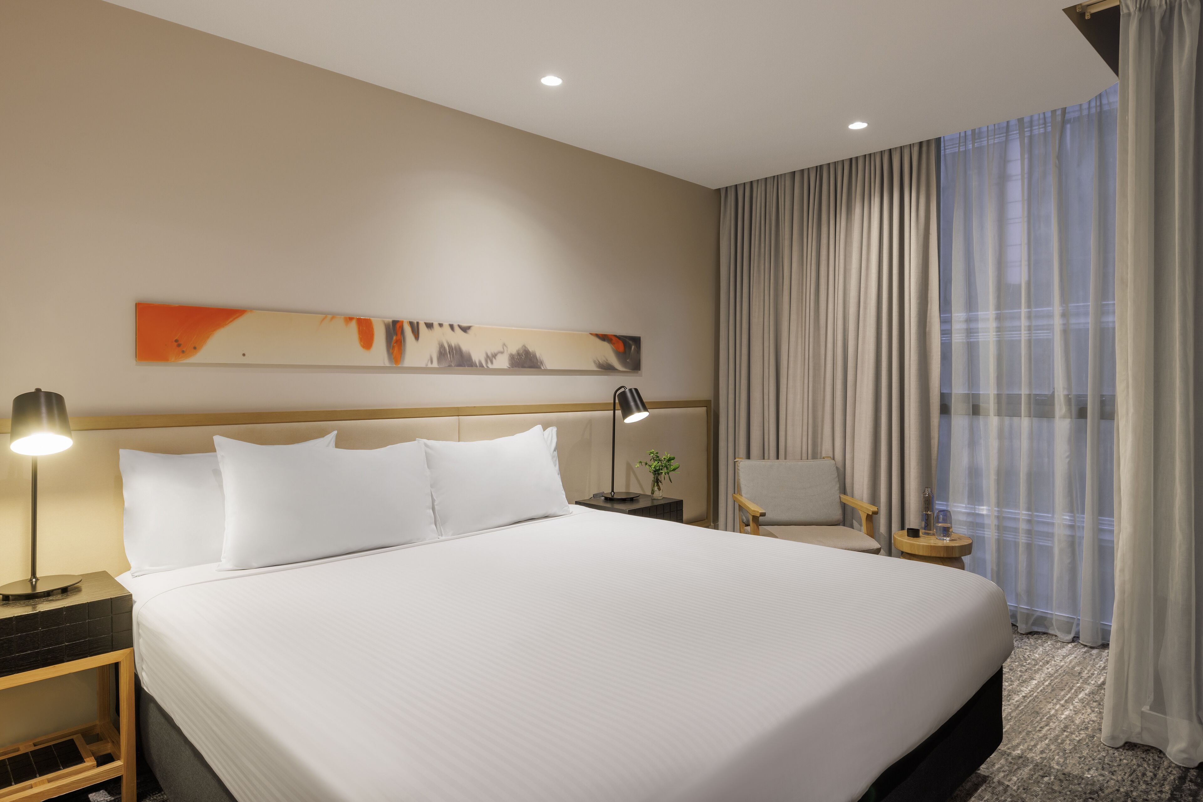 Quarto de hotel com uma cama grande, mesa de cabeceira com abajur, uma cadeira e janela com cortinas.