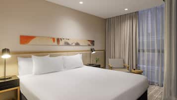 Quarto de hotel com uma cama grande, mesa de cabeceira com abajur, uma cadeira e janela com cortinas.