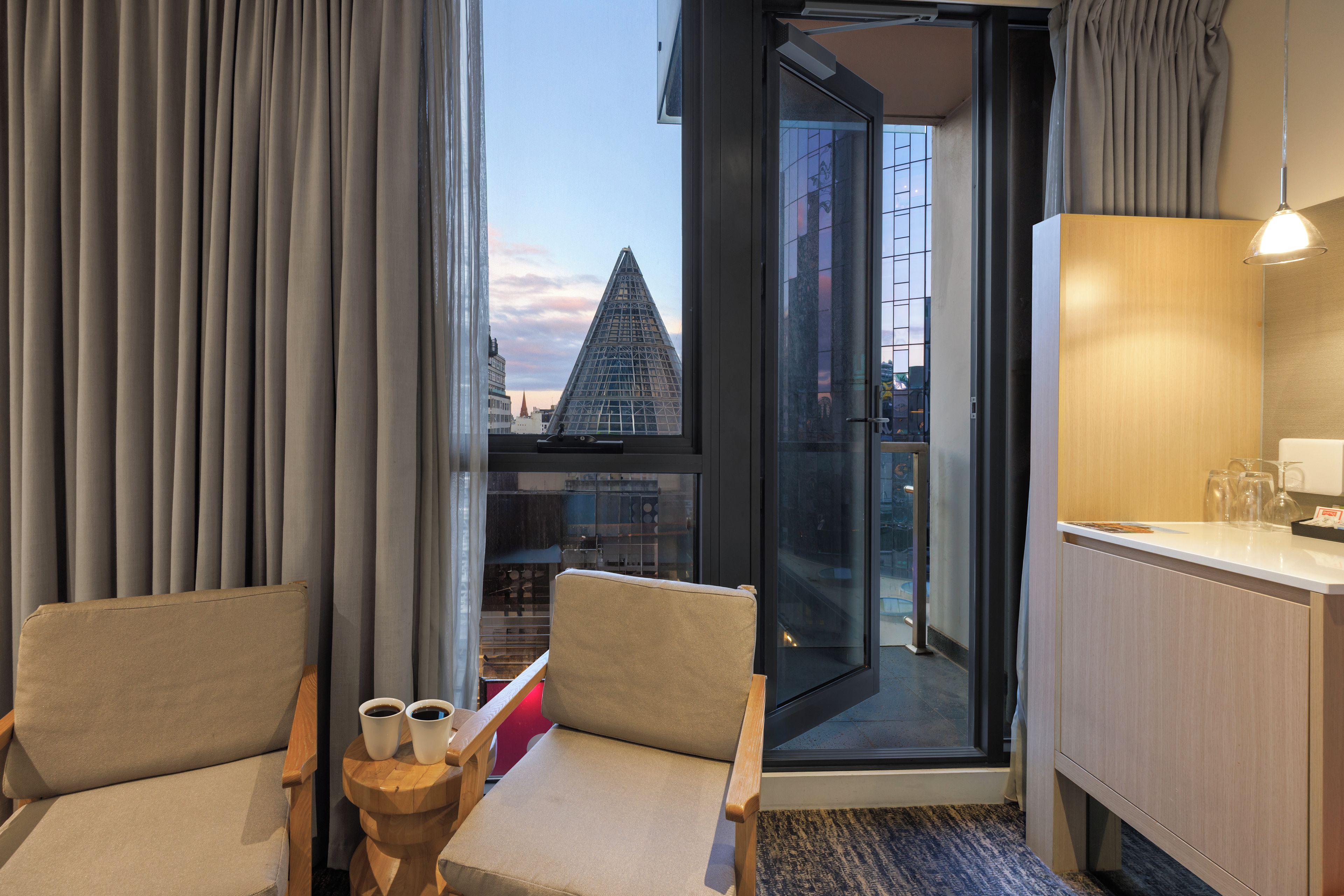 Hotelkamer met uitzicht op een modern gebouw, een houten bureau, twee stoelen en een kleine tafel met kopjes.