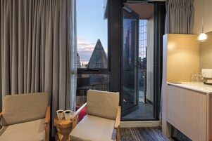 Hotellihuone, josta on näkymä moderniin rakennukseen. Huoneessa on puinen työpöytä, kaksi tuolia ja pieni pöytä, jonka päällä on kuppeja.