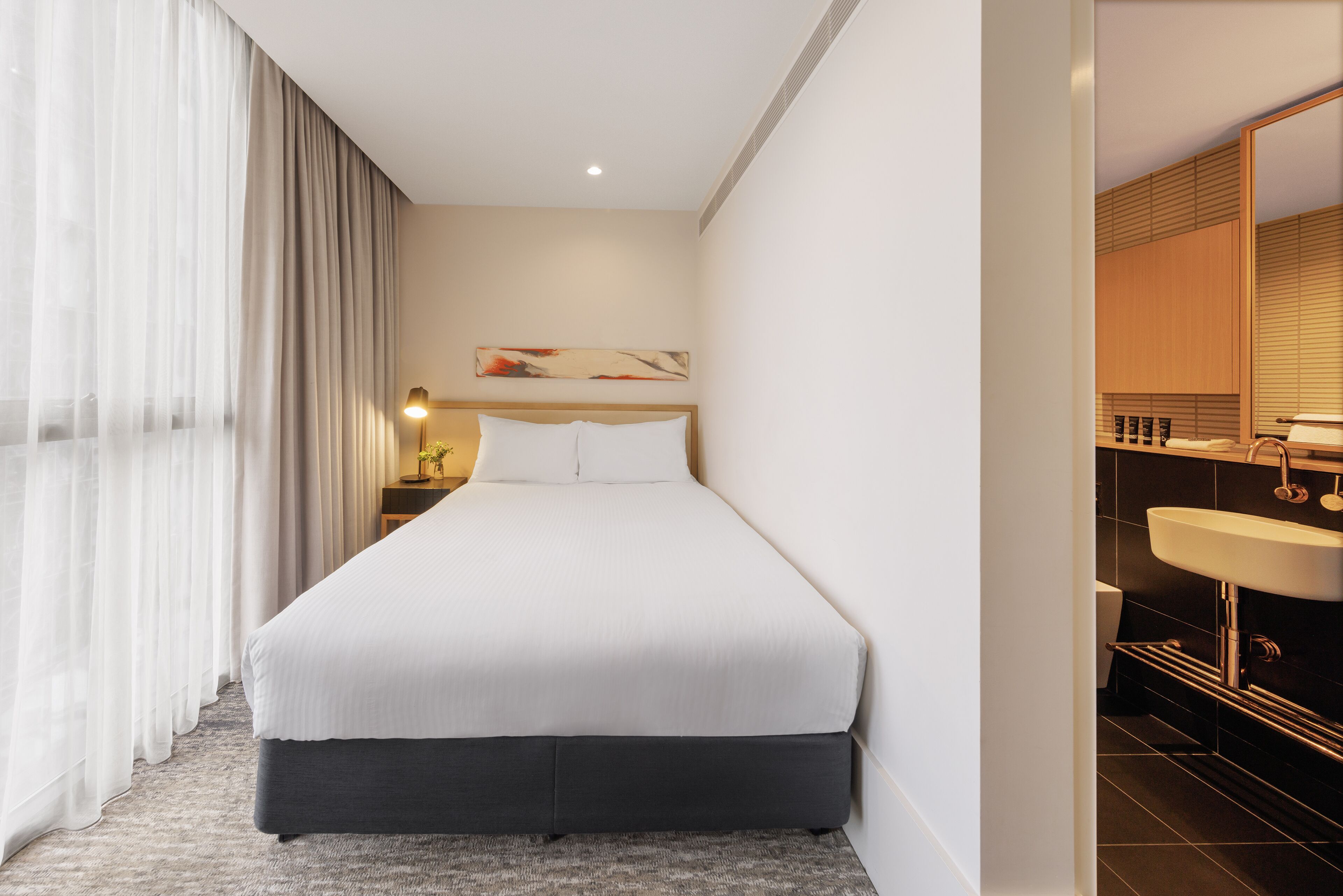 Et moderne hotelværelse med en stor seng, et natbord med lampe, et badeværelse med vask og spejl, samt udsigt over byen gennem vinduet.