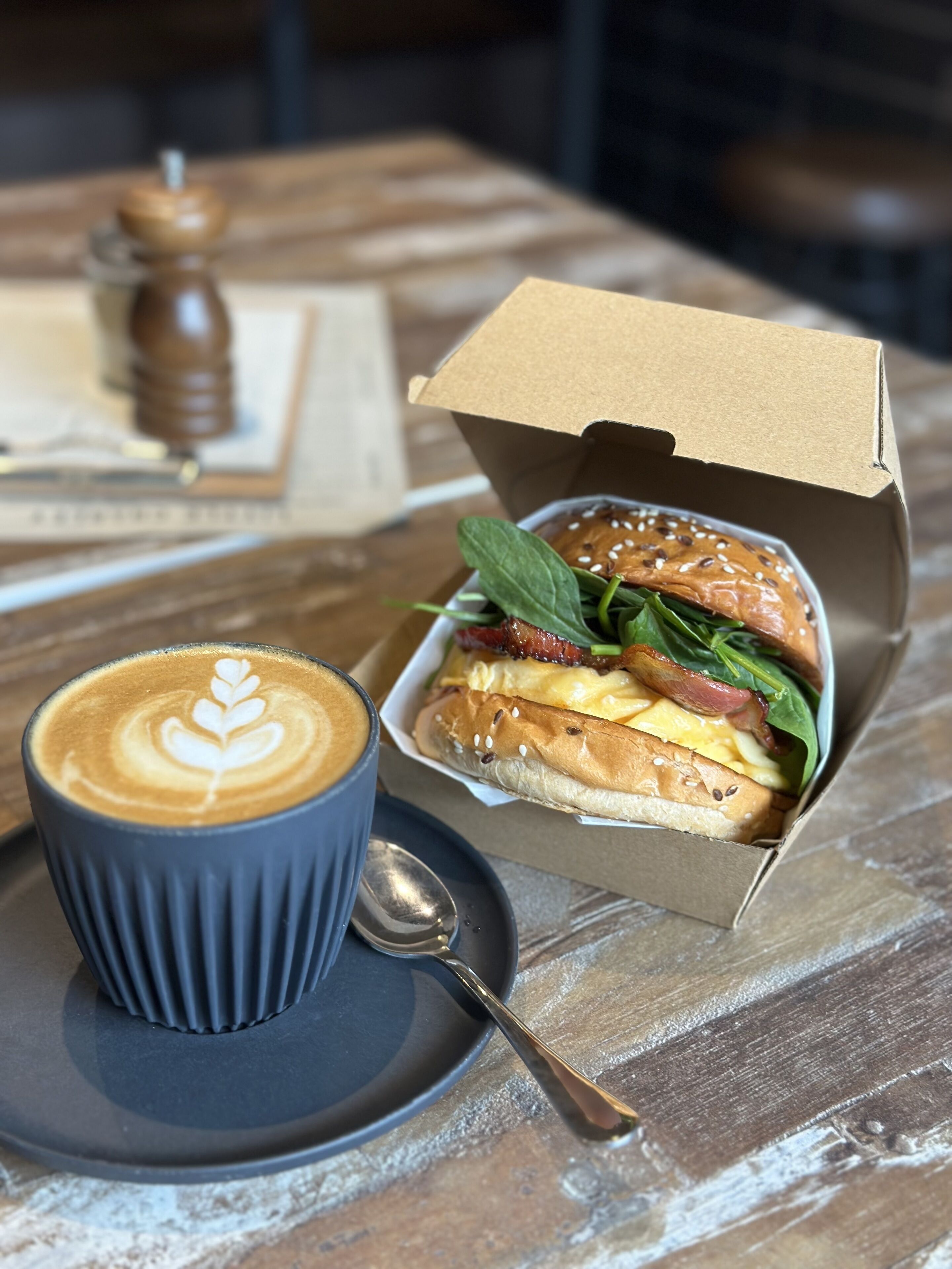 A cup of coffee with latte art on a saucer, a sandwich in a cardboard box, and a pepper grinder on a wooden table.