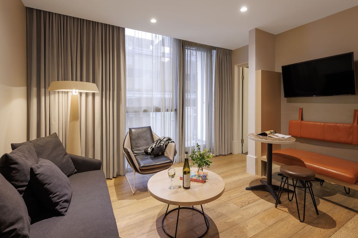 Una sala moderna con un sofá, una silla, una mesita con una botella y un vaso, y un televisor montado en la pared.
