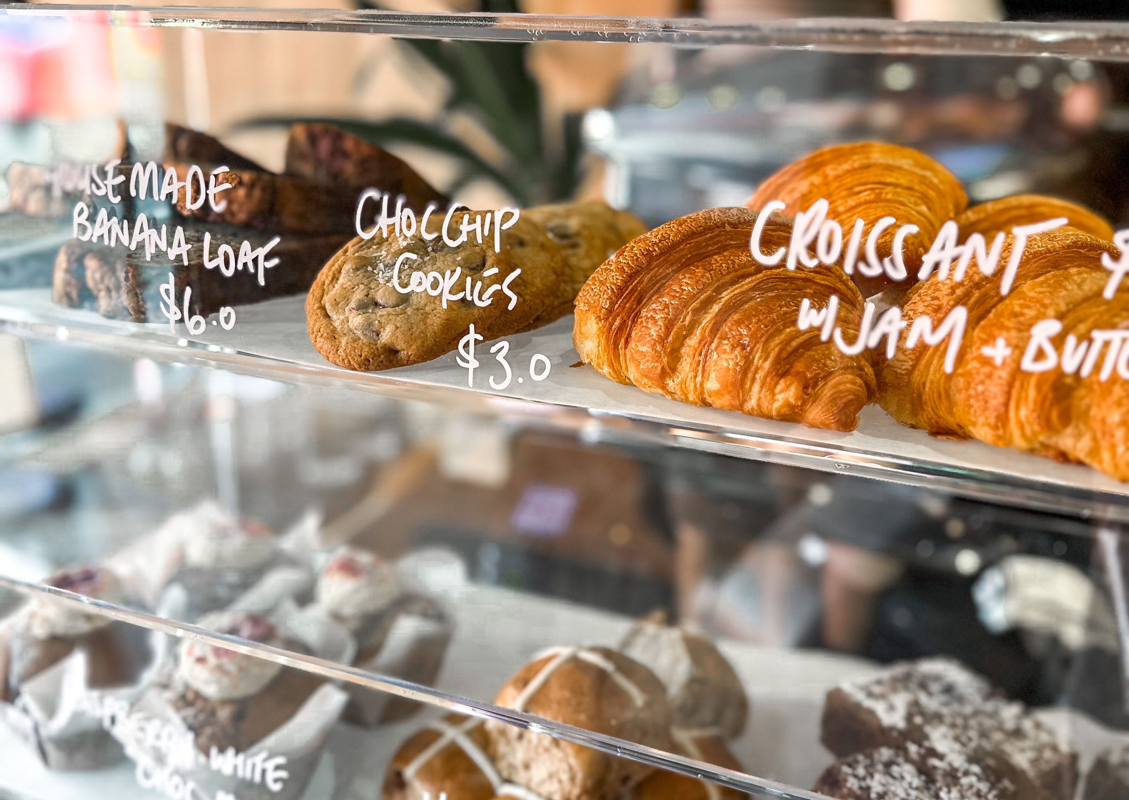A bakery display with various baked goods including banana loaf, chocolate chip cookies, croissant, and jam + butter croissant. Prices are visible.