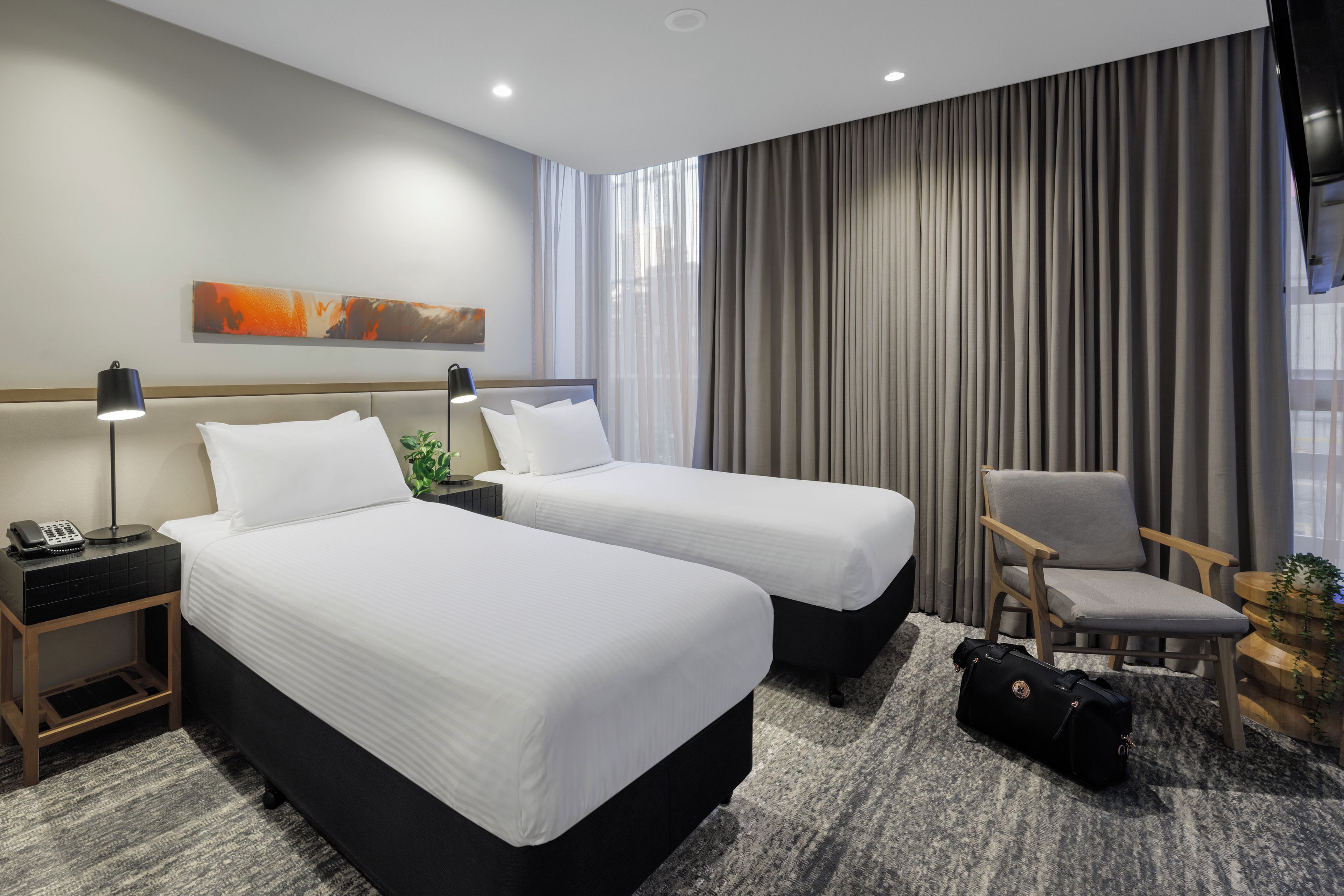 A hotel room with two beds, a desk with a lamp, a chair, and a large window with curtains.