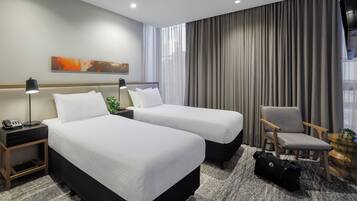A hotel room with two beds, a desk with a lamp, a chair, and a large window with curtains.