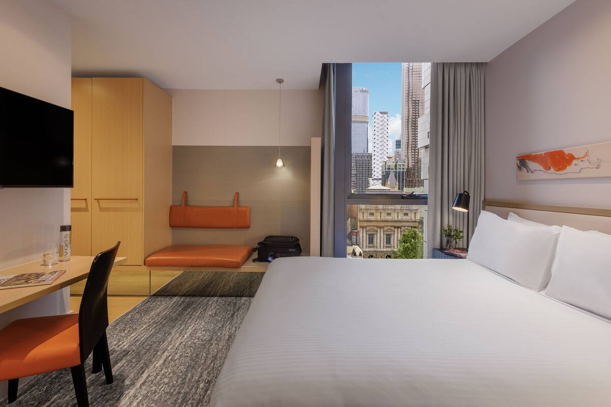 Habitación de hotel con cama, escritorio, silla, televisión y vista a la ciudad.