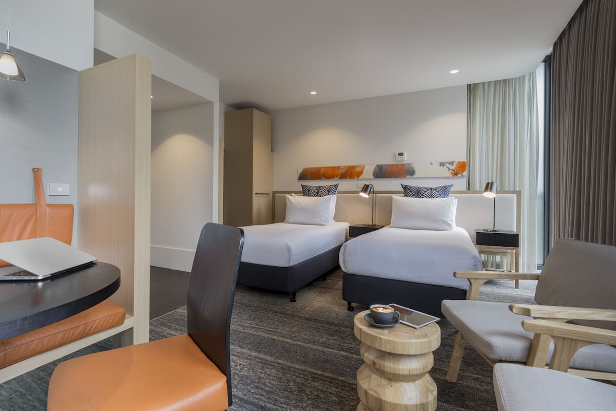 Habitación de hotel con dos camas, un escritorio con una computadora portátil, una silla y una pequeña mesa redonda con una taza.