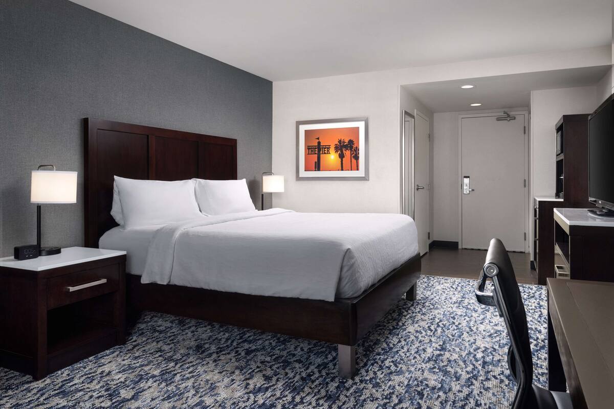 A hotel room with a large bed, two bedside tables with lamps, a desk with a chair, a TV, and a framed picture on the wall.