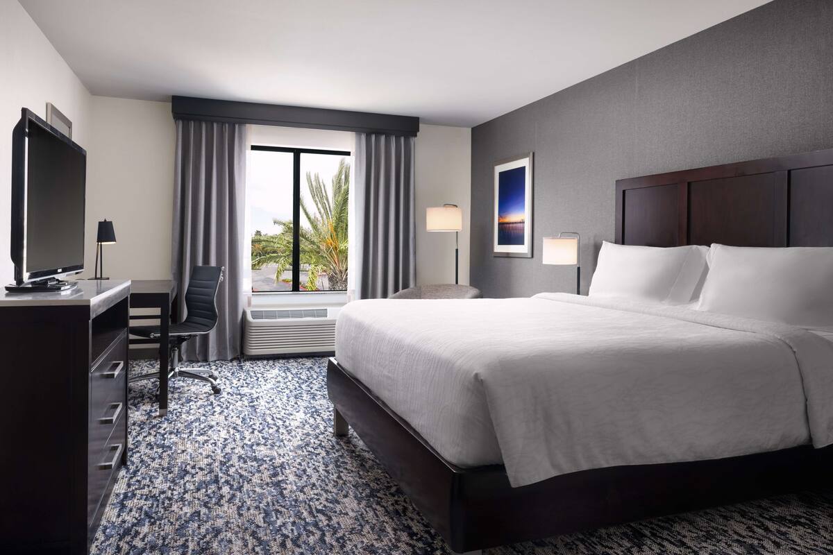 A hotel room with a large bed, a desk with a TV, a chair, a window with curtains, and a wall-mounted lamp.