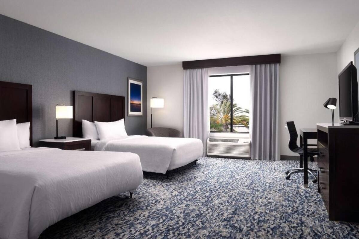A hotel room with two beds, a desk, a chair, a TV, and a window with curtains.