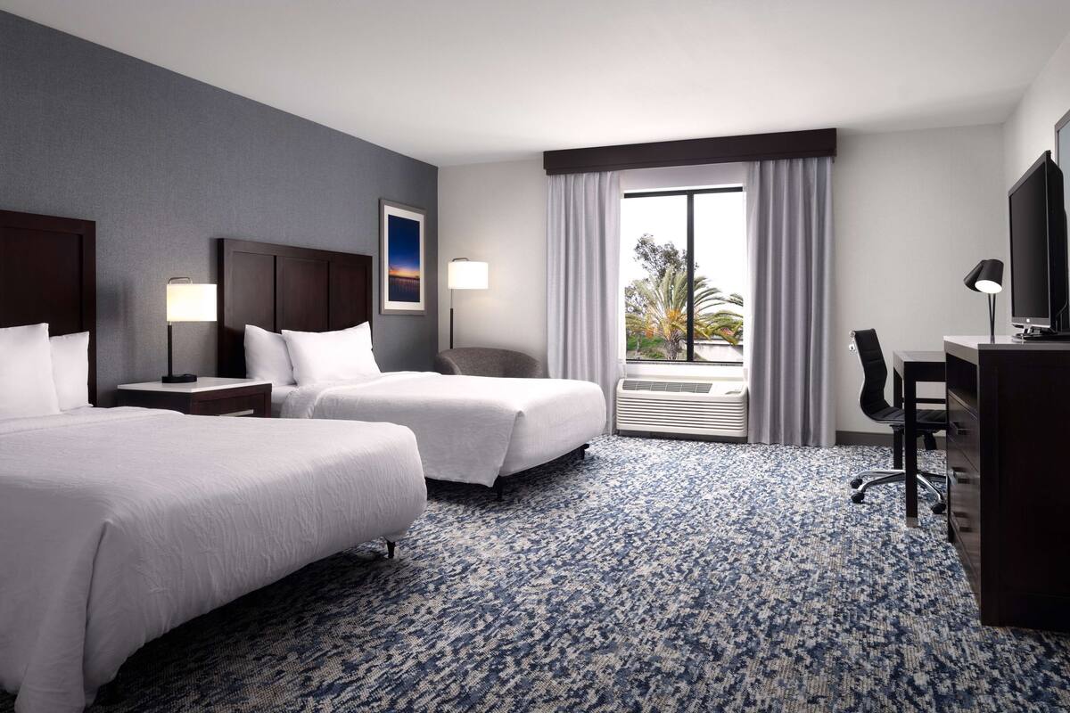 A hotel room with two beds, a desk, a chair, a television, and a window with curtains.