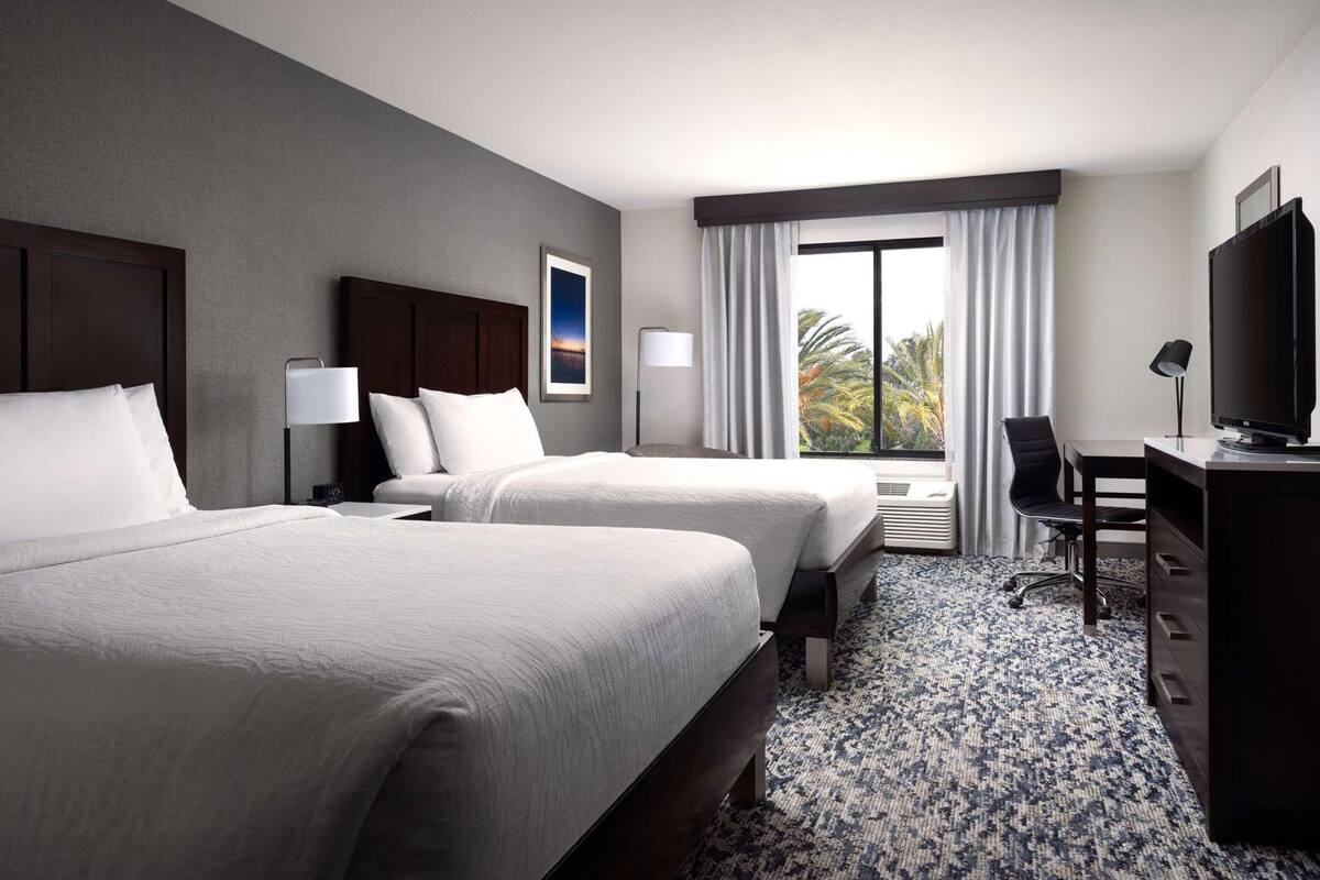 A hotel room with two beds, a desk, a TV, and a window with curtains.