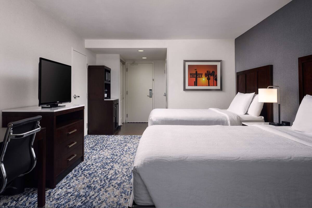 A hotel room with a bed, a desk with a TV, a chair, a wardrobe, and a painting on the wall.