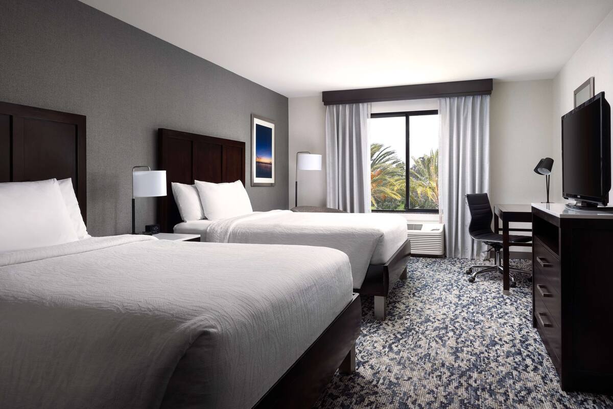 A hotel room with two beds, a desk, a television, and a window with curtains.