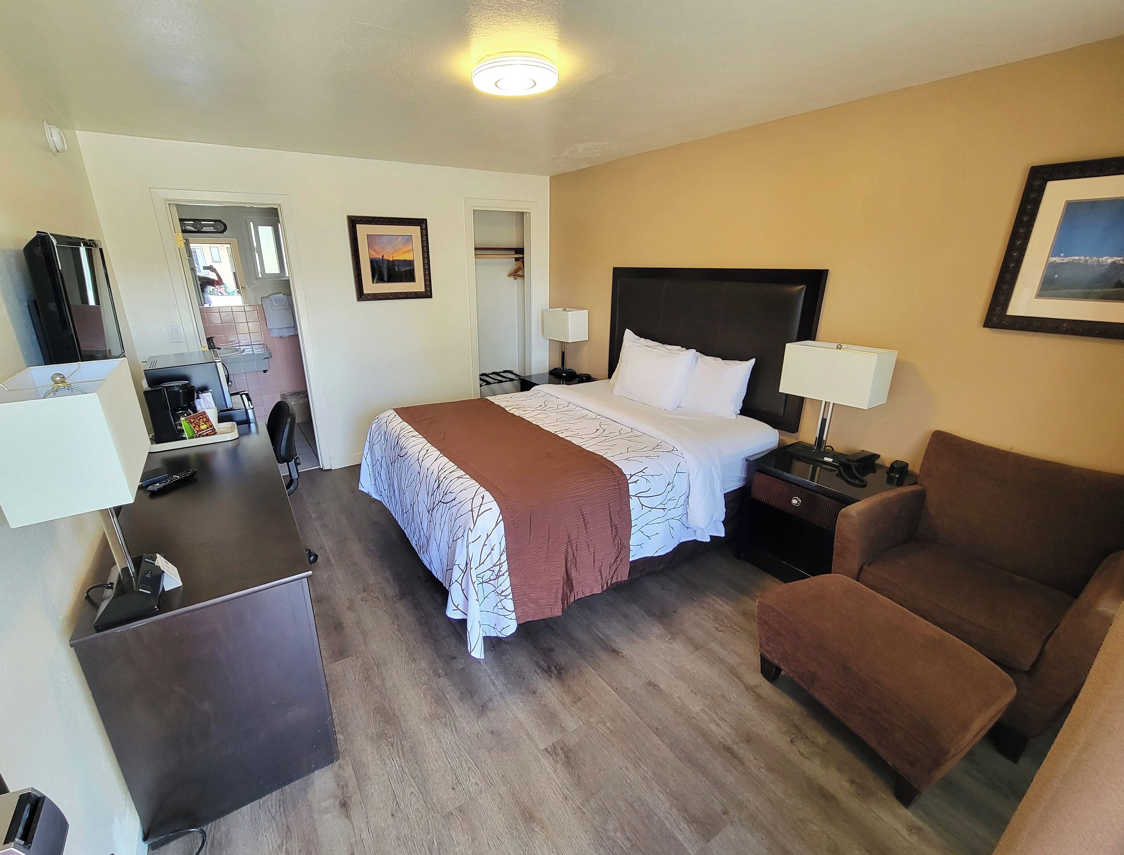 Habitación de hotel con una cama, un sillón marrón, un escritorio con lámpara, televisión y baño a la vista a través de una puerta abierta.